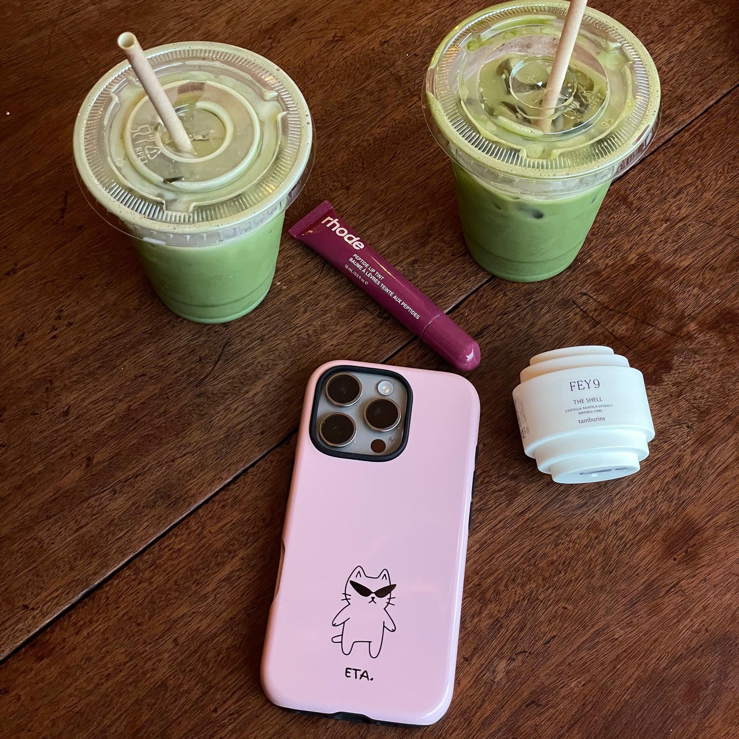 Pink phone case with a cartoon design on a wooden surface with green drinks and a lip balm.