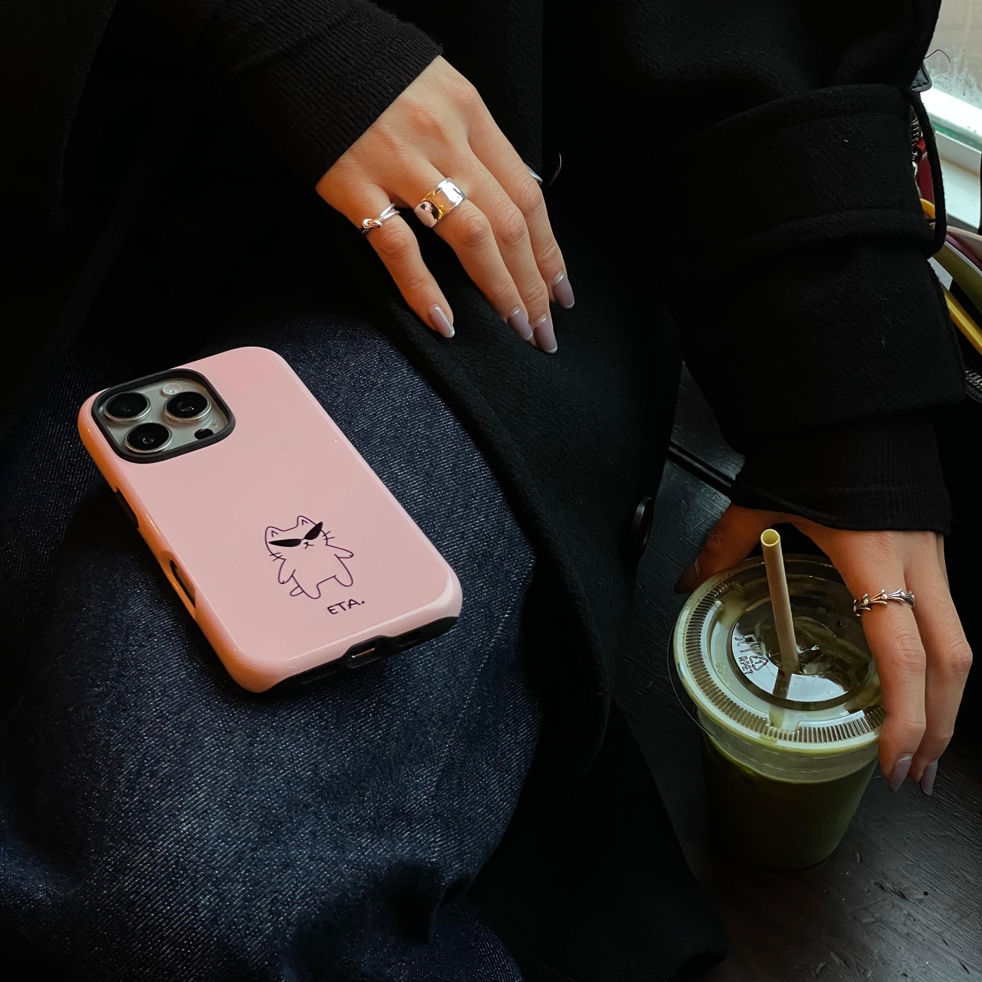 Person holding a pink phone case with a design and a green drink with a straw on a dark surface.